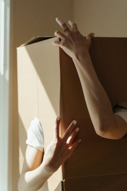 The image shows two individuals during a home relocation process, with one person lifting a large cardboard moving box, revealing part of their white sleeve, while the other person's hand, partially visible, supports the box from below. The scene takes place indoors near a doorway with natural light casting shadows on the beige wall. The box appears to be sealed with packing tape and is likely used for packing household items such as books, kitchenware, or personal belongings. The background highlights a clean, uncluttered environment, emphasizing careful handling and packing as part of a furniture transport or move. This visual aligns with professional removals services, such as those offered by Merton Removals, when preparing for house moves, packing, and loading household goods onto moving vehicles.