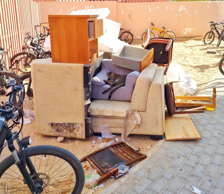 A pile of discarded household furniture and waste materials outdoors on a paved area, including an old, stained sofa with a visible patch of dirt at the bottom, a small purple armchair with a tabby cat resting on it, and various dismantled items such as wooden tables, chairs, and cabinets, some of which are overturned or broken. cardboard boxes, plastic wrapping, and packaging debris are scattered around the furniture. Bicycles are parked nearby, and a large white foam block and a brick wall with peeling paint are visible in the background. The scene suggests the process of clearing or removing bulky waste as part of a home relocation or furniture disposal service, typical of professional removals provided by Merton Removals.
