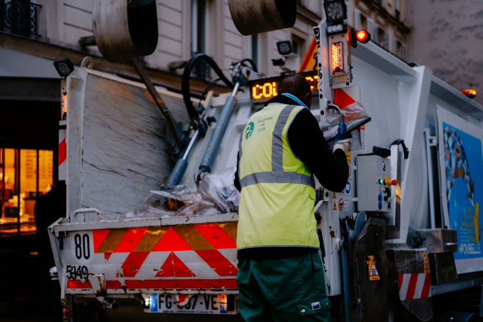 A waste collection worker wearing a high-visibility yellow vest and dark clothing is operating the controls of a large blue and white waste disposal truck parked on a residential street during evening hours. The worker is facing the truck's side panel, which features various controls, warning lights, and a digital display showing the code 'CO1'. Inside the truck's open rear container, there are stacked cardboard boxes, plastic-wrapped packages, and wooden pallets, indicating the collection of bulky waste items such as furniture and appliances. The area is illuminated by the truck's onboard lights and ambient street lighting, with a building featuring lit windows visible in the background. The scene captures the process of waste collection and logistics involved in furniture transport or home relocation services provided by Merton Removals, demonstrating an active loading or waste removal operation related to house moves and bulky waste disposal.