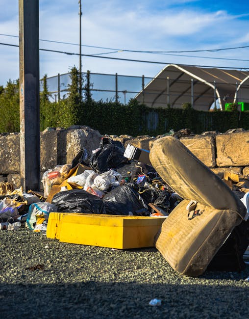 A pile of discarded household furniture and waste materials outdoors on a paved area, including an old, stained sofa with a visible patch of dirt at the bottom, a small purple armchair with a tabby cat resting on it, and various dismantled items such as wooden tables, chairs, and cabinets, some of which are overturned or broken. cardboard boxes, plastic wrapping, and packaging debris are scattered around the furniture. Bicycles are parked nearby, and a large white foam block and a brick wall with peeling paint are visible in the background. The scene suggests the process of clearing or removing bulky waste as part of a home relocation or furniture disposal service, typical of professional removals provided by Merton Removals.