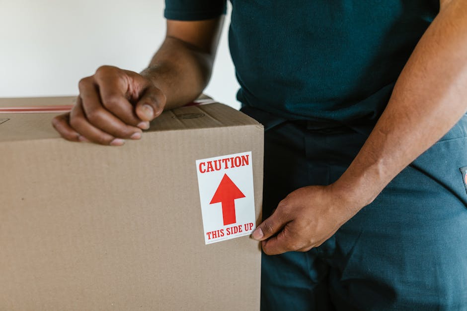 Close-up view of a person wearing a dark green T-shirt and blue trousers, carefully handling a large cardboard box during a home relocation process. The individual is holding the box with both hands, with one hand gripping a corner and the other supporting the side, while standing inside a room with neutral-colored walls. The box features a prominent red and white caution label that reads 'CAUTION THIS SIDE UP' with an upward-pointing arrow, indicating proper orientation for safe handling. In the background, part of a door frame and a light-colored interior wall are visible, suggesting the loading or unloading phase of furniture transport. The scene captures the careful packing and moving of household items, with an emphasis on safety and proper handling during furniture transport or packing and moving services provided by Merton Removals, who specialise in house removals and relocation logistics.