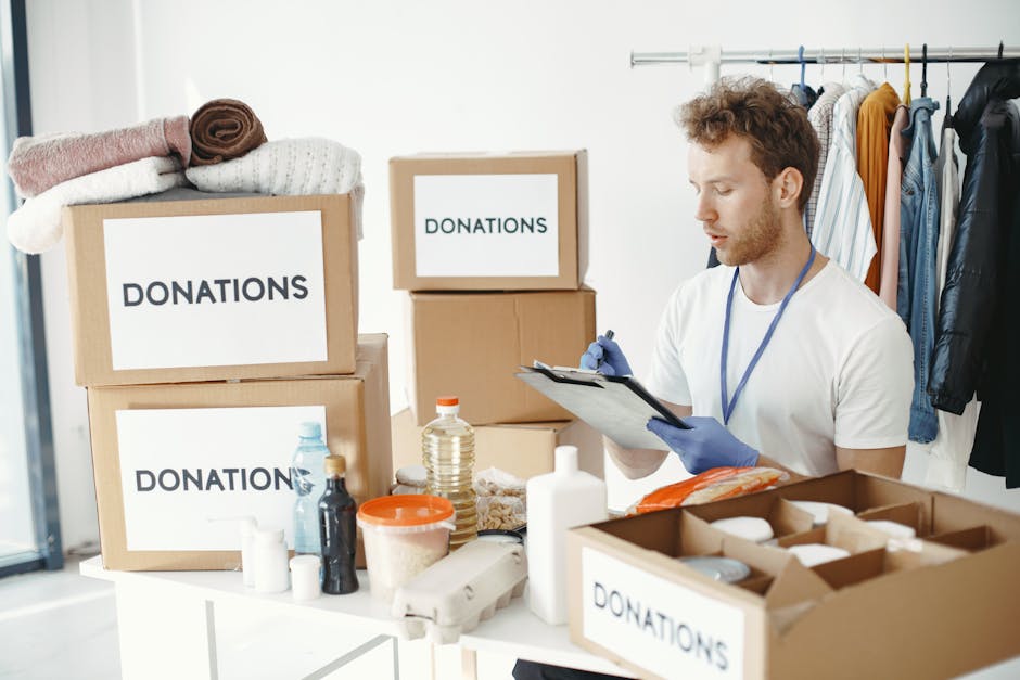 Close-up view of a person wearing a dark green T-shirt and blue trousers, carefully handling a large cardboard box during a home relocation process. The individual is holding the box with both hands, with one hand gripping a corner and the other supporting the side, while standing inside a room with neutral-colored walls. The box features a prominent red and white caution label that reads 'CAUTION THIS SIDE UP' with an upward-pointing arrow, indicating proper orientation for safe handling. In the background, part of a door frame and a light-colored interior wall are visible, suggesting the loading or unloading phase of furniture transport. The scene captures the careful packing and moving of household items, with an emphasis on safety and proper handling during furniture transport or packing and moving services provided by Merton Removals, who specialise in house removals and relocation logistics.