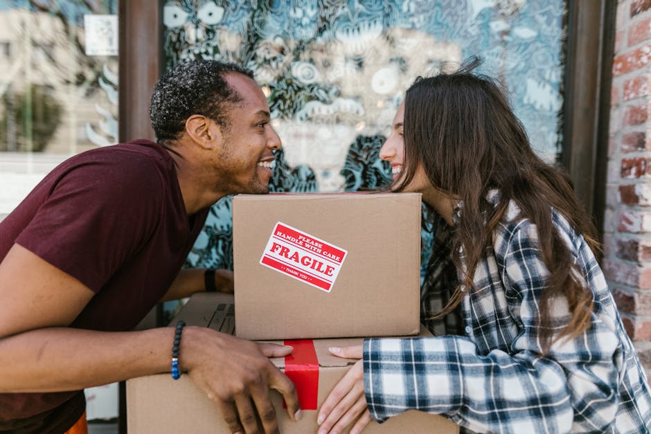 Two individuals engaged in a home relocation process outdoors in front of a building with large windows and brick walls. The man, with short, curly hair and wearing a maroon t-shirt, is smiling and leaning forward as he holds a cardboard box labeled 'Fragile' with red tape, indicating careful handling. The woman, with long, wavy brown hair and dressed in a black-and-white plaid shirt, is also smiling and appears to be in the process of handing or receiving the box. Additional packing materials, such as cardboard boxes and protective wrapping, are visible, as well as a moving trolley nearby in the outdoor space. Natural lighting illuminates the scene, capturing a moment of coordination during furniture transport or packing for a move, supported by [COMPANY_NAME], a professional removals service specializing in house removals and relocation logistics.