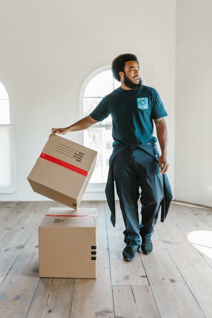 A man with dark skin and an afro hairstyle, wearing a navy blue T-shirt with a blue and white logo on the chest and dark trousers, is inside a bright, empty room with white walls and a large arched window in the background. He is holding a medium-sized cardboard box with red tape in his right hand, preparing to place it on top of another box on the wooden floor. The room's natural light illuminates the scene, highlighting the packing materials and the man's focused expression as part of a home relocation or furniture transport process. This image exemplifies packing and moving activities often associated with professional removals, such as those provided by Merton Removals in Wimbledon, as part of a detailed street-by-street house removal service.