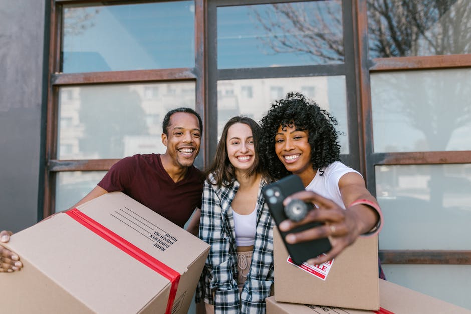 Two individuals engaged in a home relocation process outdoors in front of a building with large windows and brick walls. The man, with short, curly hair and wearing a maroon t-shirt, is smiling and leaning forward as he holds a cardboard box labeled 'Fragile' with red tape, indicating careful handling. The woman, with long, wavy brown hair and dressed in a black-and-white plaid shirt, is also smiling and appears to be in the process of handing or receiving the box. Additional packing materials, such as cardboard boxes and protective wrapping, are visible, as well as a moving trolley nearby in the outdoor space. Natural lighting illuminates the scene, capturing a moment of coordination during furniture transport or packing for a move, supported by [COMPANY_NAME], a professional removals service specializing in house removals and relocation logistics.