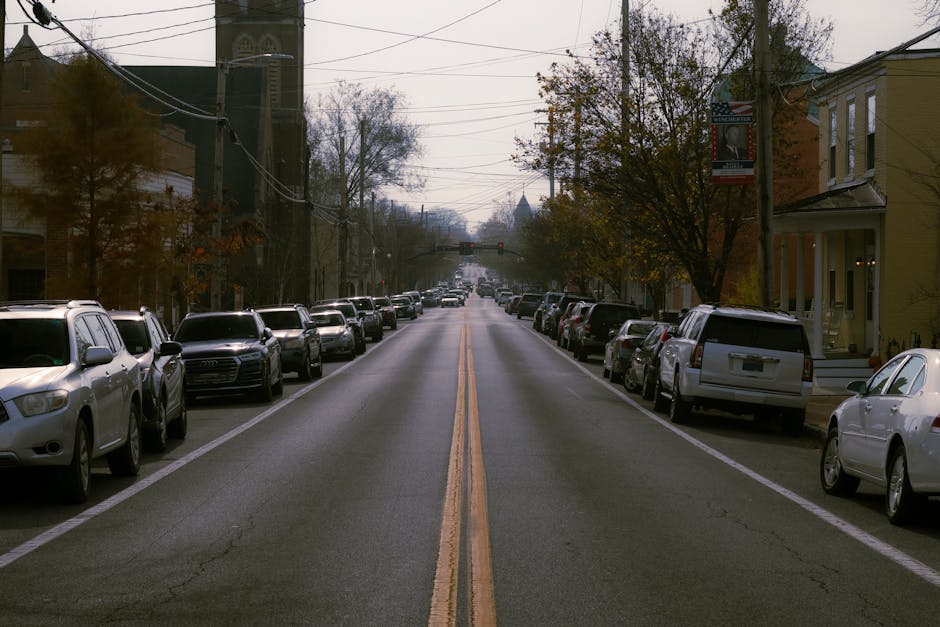 A straight residential street in Merton with parked cars lining both sides of the road, including sedans, SUVs, and hatchbacks in various colours such as white, silver, and black. The street surface is paved with asphalt, featuring a central double yellow line indicating no overtaking. Overhead, multiple power lines stretch across the street between utility poles. On the right side, there are houses with facades partially visible, some with front gardens or steps leading to the front doors. Autumn trees with sparse foliage are visible along the sidewalks, with some leaves scattered on the pavement. In the distance, the road appears to ascend slightly, with a faint view of more buildings and possibly church spires or distinctive rooftops in the background. This scene depicts a typical neighbourhood maintained during daylight hours, with natural light and overcast skies, and relates to home relocation or furniture transport logistics, as handled by companies like Merton Removals.