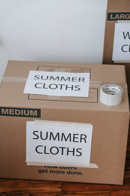 A close-up view of a cardboard moving box labeled 'SUMMER CLOTHS' with a smaller piece of paper reading 'SUMMER CLOTHS' taped to the front. The box is positioned on a wooden floor and appears to be part of a home packing process for a house relocation. Inside the box, visible packing materials include a roll of white packing tape with black text, and the box is marked with a black label indicating 'MEDIUM' size. In the background, another larger box or parcel is partially visible, also labeled with service-related text, suggesting the context of professional removals by Merton Removals. The setting indicates organized packing efforts for a move, with furniture and appliance transport preparations likely underway as part of a comprehensive packing and moving process on Kingston Road, Merton.