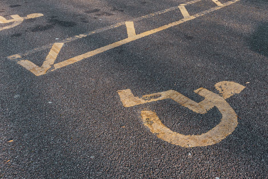 Close-up of a designated accessible parking space on an asphalt surface, featuring a yellow wheelchair symbol painted on the ground with some signs of wear and peeling. The space includes white diagonal lines at the top edge, indicating accessible parking zone boundaries. This parking area is located outdoors, adjacent to a building entrance or loading zone, relevant for home relocation or furniture transport logistics. The photograph emphasizes the importance of accessible parking for residential removals, as managed by Merton Removals, especially when planning moves near Wimbledon Common, with considerations for parking access and loading areas during the loading process.