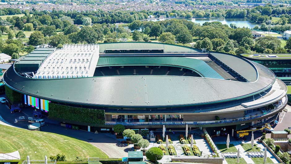 An aerial view of a large oval-shaped building with a green metallic roof, situated in a lush green landscape with trees and grass surrounding it. The building appears to be a sports stadium or arena, with an open central area and a partially covered seating section. Nearby, there are vehicles parked along a driveway and a paved area with outdoor furniture, including tables with large umbrellas, indicating outdoor seating or event space. The surrounding environment includes trees, a small lake in the distance, and rural or parkland scenery. The image showcases the building's modern architectural design and its context within a spacious, green environment, relevant for house removals or relocation services such as those offered by Merton Removals near Wimbledon Common, providing insight into the accessibility and parking considerations for large property moves or furniture transport.