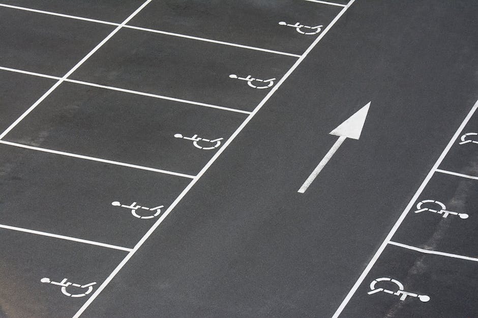 Close-up of a designated accessible parking space on an asphalt surface, featuring a yellow wheelchair symbol painted on the ground with some signs of wear and peeling. The space includes white diagonal lines at the top edge, indicating accessible parking zone boundaries. This parking area is located outdoors, adjacent to a building entrance or loading zone, relevant for home relocation or furniture transport logistics. The photograph emphasizes the importance of accessible parking for residential removals, as managed by Merton Removals, especially when planning moves near Wimbledon Common, with considerations for parking access and loading areas during the loading process.