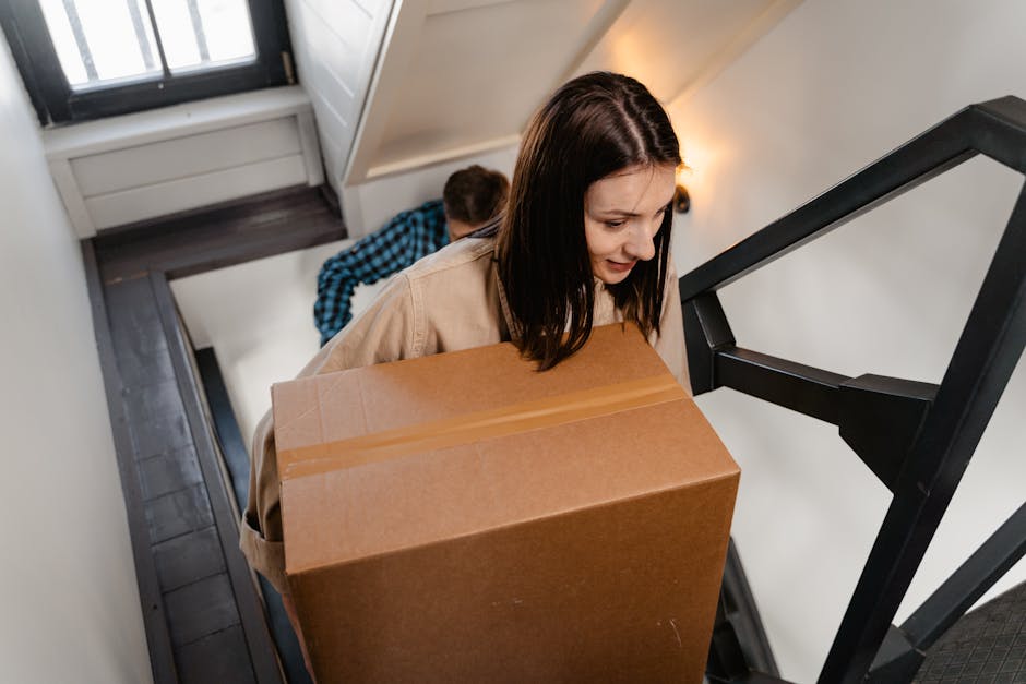 A woman with dark brown hair, wearing a beige coat, is carrying a large rectangular cardboard box up a narrow staircase in a residential property. Behind her, a man wearing a blue and black checked shirt is also moving items, slightly blurred due to the focus on the woman. The staircase has white walls and a black metal railing, with a wooden door at the top. The steps are made of dark wood, and the area is lit with warm lighting. The woman is focused on the task, carefully navigating the stairs during a home relocation process. This scene illustrates furniture transport and packing during a house move, with visible cardboard packing materials and the use of proper lifting techniques, supported by a professional removals service such as Merton Removals, which manages secure stair-only moves in South Wimbledon.