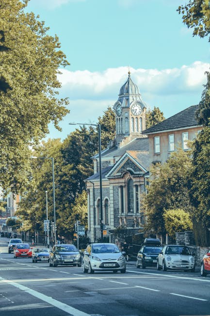A wide view of a street in SW19 showing several parked cars along the curb, with some in motion, on a sunny day with a clear blue sky. Prominent in the background is a historic building with a clock tower featuring a domed roof, decorative stonework, and arched windows, surrounded by lush green trees that partially shade the street. In the foreground, a partially visible moving van with the rear doors open is positioned on the pavement, with packing supplies such as cardboard boxes, plastic wrapping, and furniture blankets visible inside and around the loading area. A hand truck or trolley appears to be used for transporting boxes or furniture. The scene captures the typical environment of home relocation or furniture transport, with the presence of a professional removals company ensuring the safe loading and unloading of items during a house move. Such settings are common when coordinating moving logistics in urban areas like SW19, especially where street parking suspensions for moves may be relevant, as highlighted in the page about street parking permits on mertonremovals.co.uk.