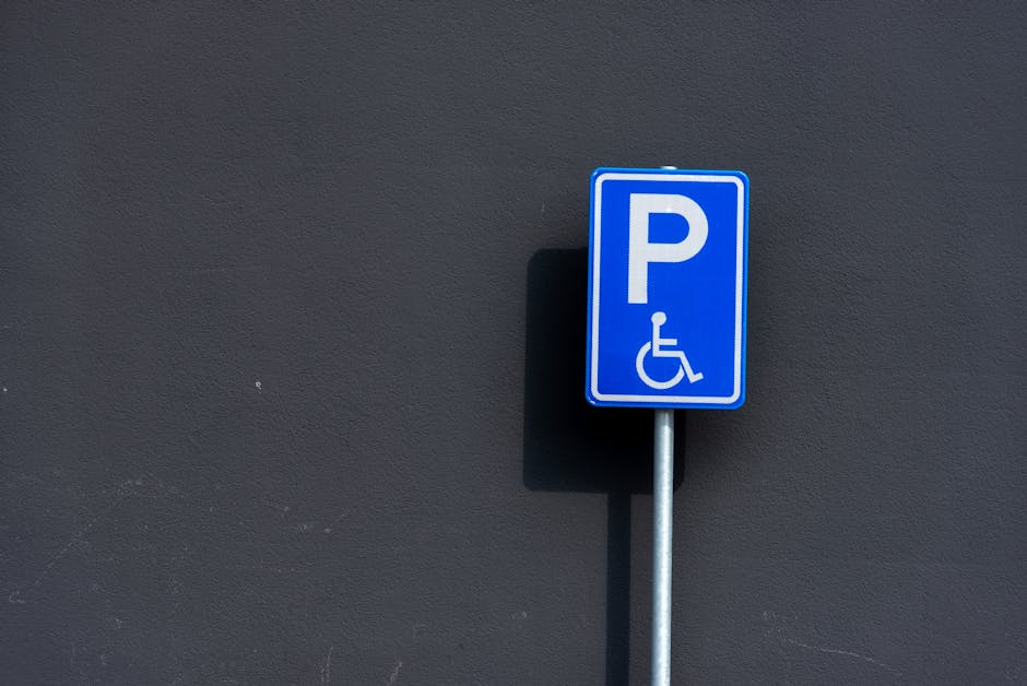 A close-up image of a blue and white parking sign with a large letter 'P' and a wheelchair symbol, mounted on a metal post against a dark grey textured wall. The sign indicates designated parking for disabled individuals and is positioned outdoors, likely on a street or in a parking area. The lighting is natural, casting a shadow of the sign onto the wall. This parking sign is relevant to city regulations and permits required for house removals or home relocation services, such as those provided by Merton Removals, especially when coordinating parking suspensions and permits for moving houses in the SW19 area. The image underscores the importance of understanding parking restrictions and permits when planning efficient furniture transport and moving logistics.